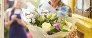 Smiling woman receiving a beautiful bouquet of fresh flowers with white roses, lilies, and purple blossoms at her doorstep, with a florist in the background taking notes.